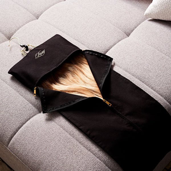 Light brown hair extensions in a black Luxy-branded case with gold zipper pulls, elegantly displayed on a grey textured couch.