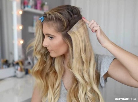 A woman gracefully installing blonde tape-in extensions into her hair, which is pinned with a clip.