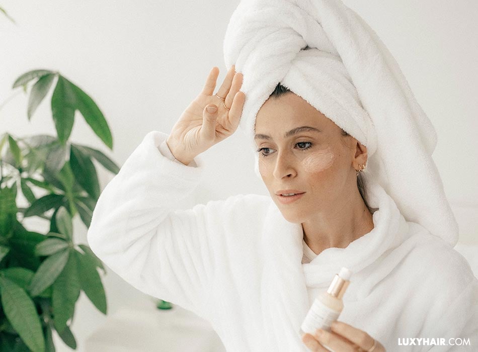 A woman in a bathrobe holding a bottle and applying creame on her face.