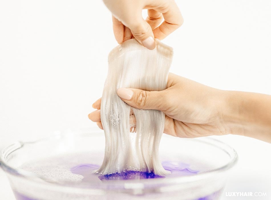 Blonde tape-in extensions are being pulled from a bowl of purple shampoo mixed with water.