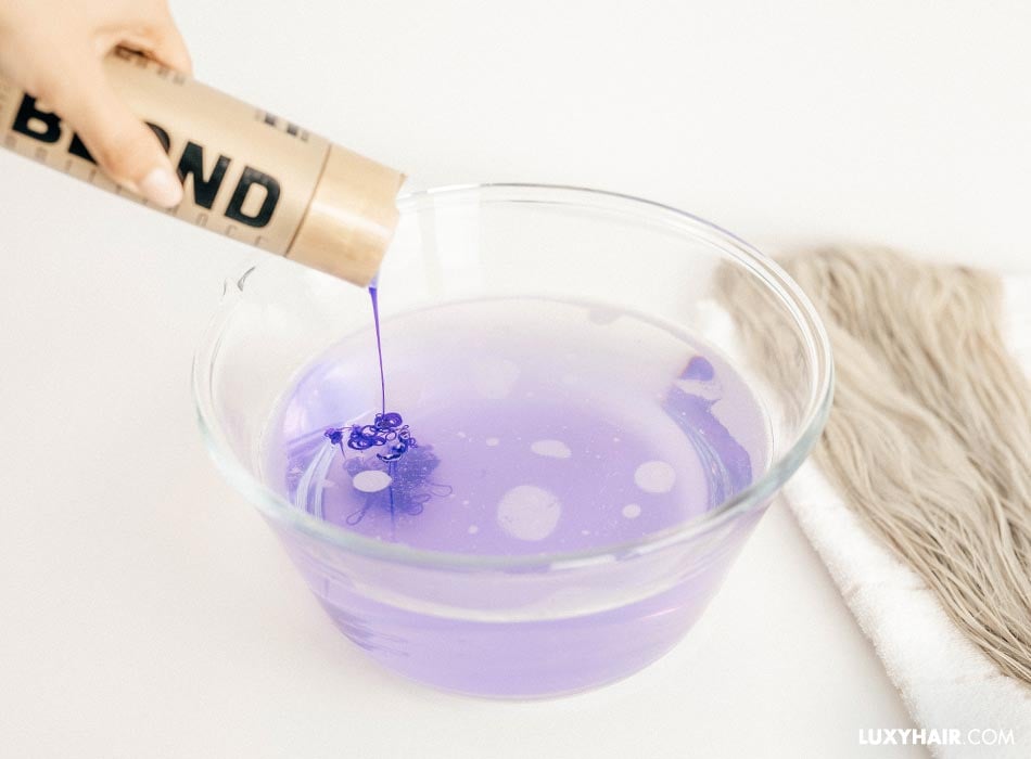 Purple shampoo being poured into a bowl of water with blonde hair extensions.