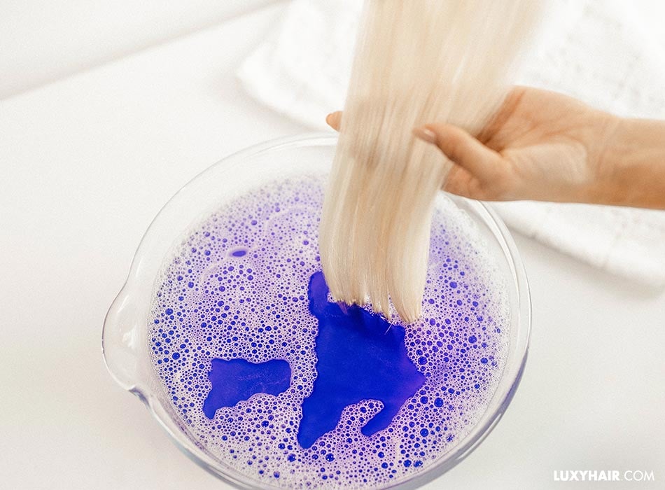 Blonde tape-in extensions are being put into a bowl of purple shampoo mixed with water.