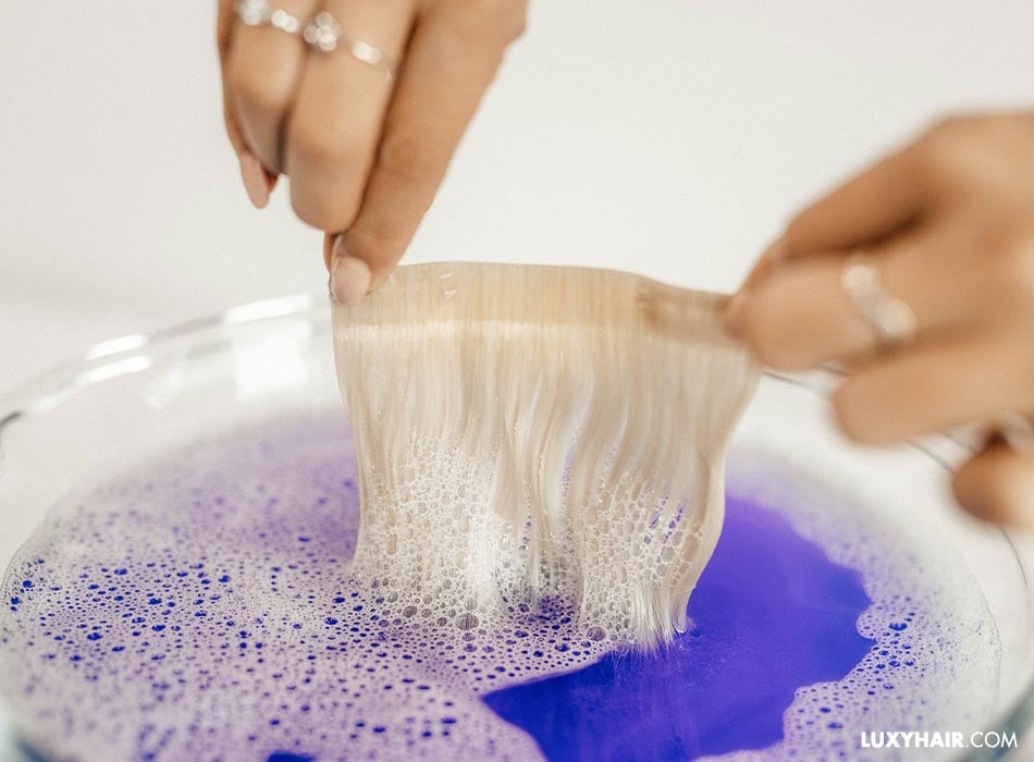 Blonde tape-in extensions are being pulled out of a bowl of purple shampoo mixed with water.