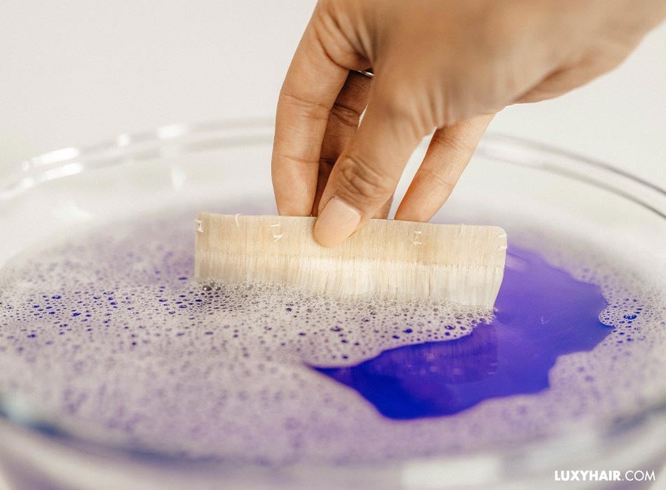 Blonde tape-in extensions are being washed in a bowl with water and purple shampoo.